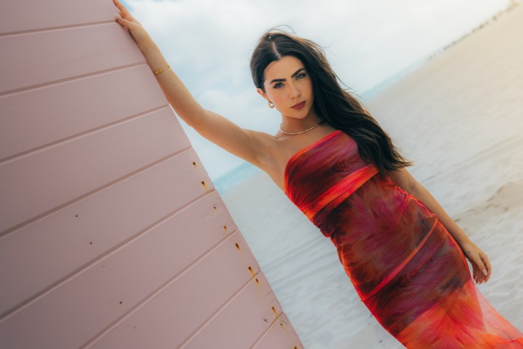 Jade Picon apoiada em parede bege usando vestido tomara que caia vermelho em tule com detalhes coloridos. 