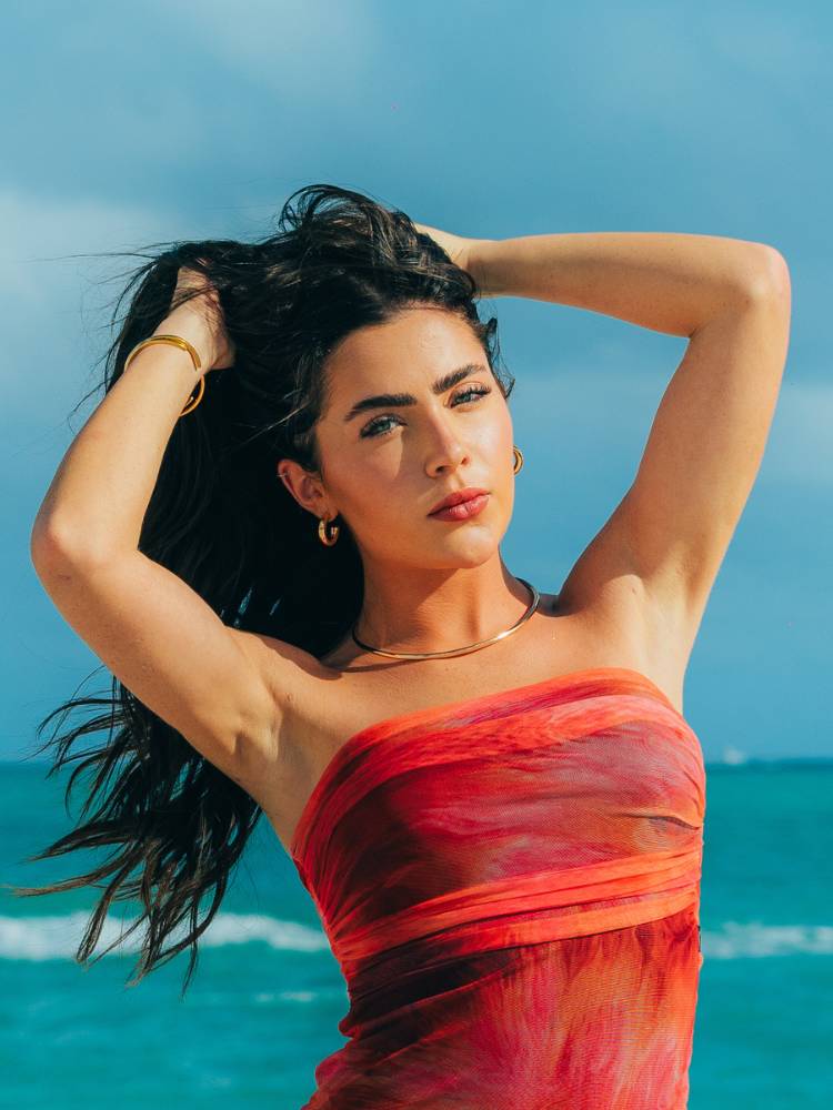 Jade Picon na praia segurando o cabelo e usando o vestido tomara que caia vermelho em tule. 