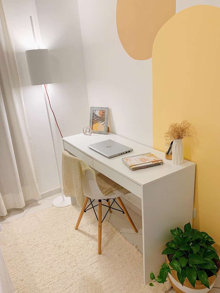 Home office em tons de branco e laranja, com mesa branca, computador, tapete e cestinho com jiboia. 
