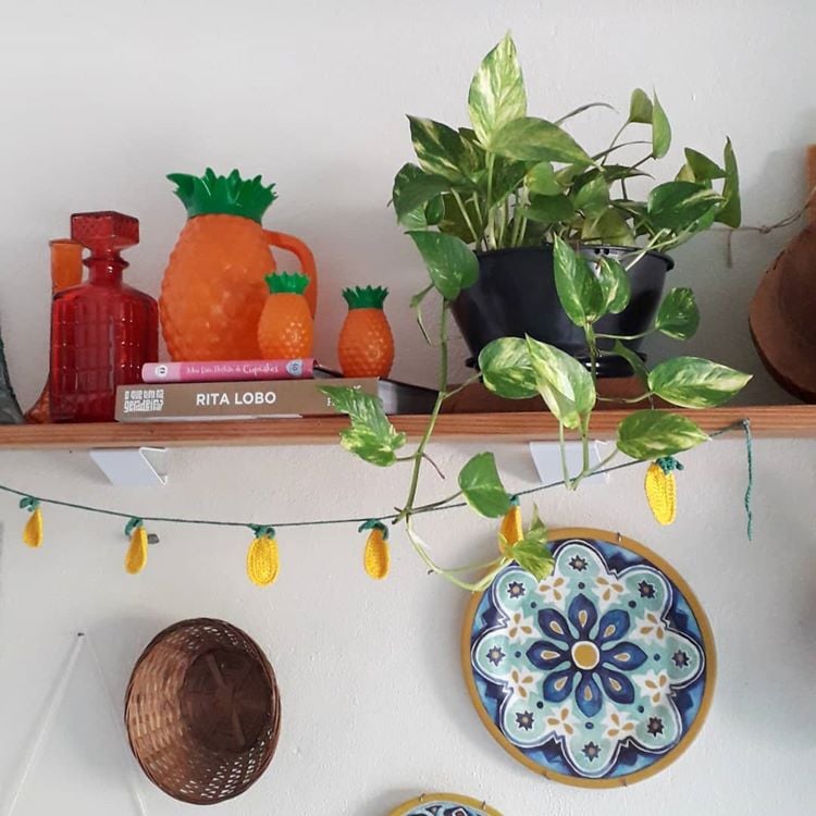 Prateleira de madeira com livros, jarra de abacaxi, vaso e jiboia pendente. 