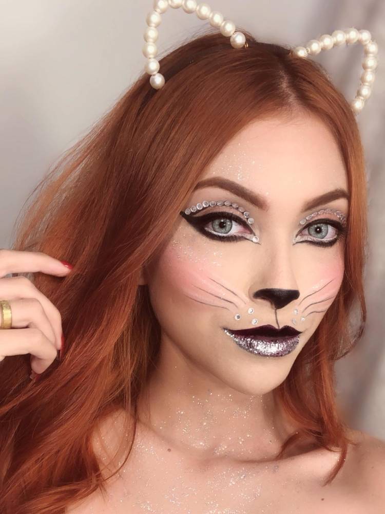 Mulher de pele clara e cabelo ruivo usando maquiagem de gato com tiara e brilho. 