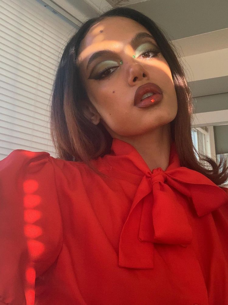 Mulher de pele morena usando camisa vermelha, sombra verde, delineado verde escuro e strass vermelhos e verdes