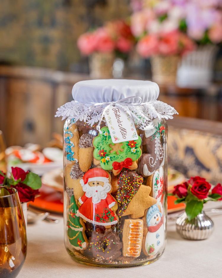 Pote de vidro com biscoitos caseiros temáticos de Natal em mesa posta com pequenos arranjos de rosas vermelhas