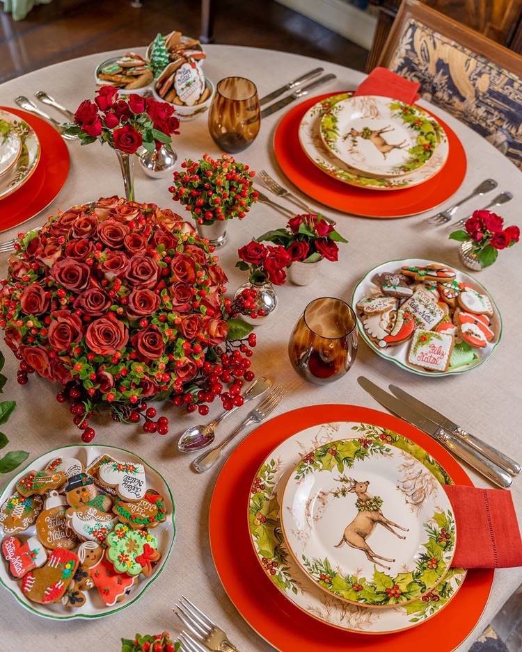 Linda mesa de Natal decorada com biscoitos caseiros decorados com frases e desenhos típicos de Natal. Os pratos são ilustrados com desenho de rena e plantas. Na mesa posta há arranjos de rosas vermelhas falsas.