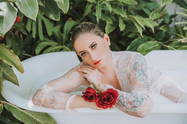 mulher loira com roupas claras e molhadas em uma banheira na natureza segurando rosas vermelhas usadas em simpatias para o amor