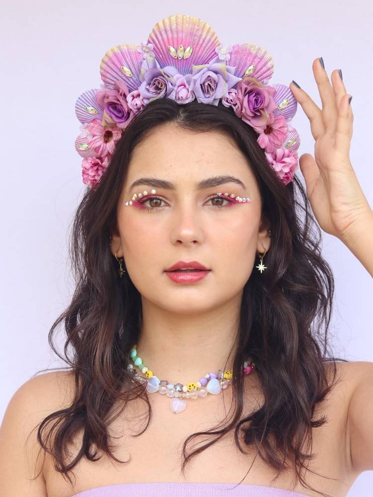 Mulher de pele clara e cabelo escuro usando tiara de concha e flores em tons de lilás e rosa. 