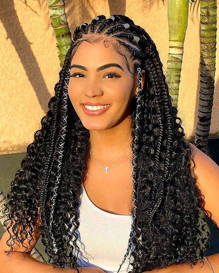 Trança nagô e box braid eem cabelo cacheado grande, com fio de seda branco e joias. Mulher usa regata branca e está em área com pé de palmeira 
