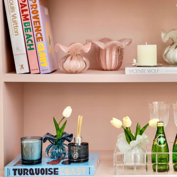 Estante rosa com vaso de planta, vela branca redonda, vasos rosas e livros. 