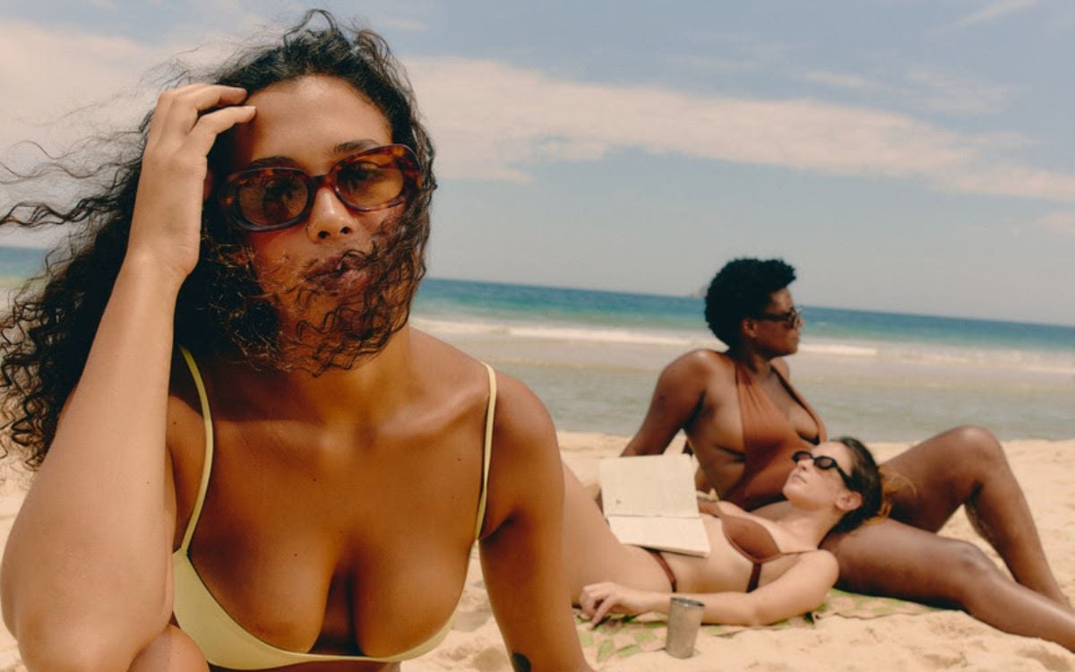 foto de três mulheres deitadas na areia da praia tomando sol, usando óculos de sol vintage e biquínis em tons terrosos