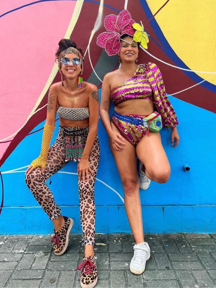 duas amigas usando tendências de fantasia de Carnaval 2024: uma usa calça de oncinha e a outra tem tiara de flor maximalista e conjunto de lantejoulas