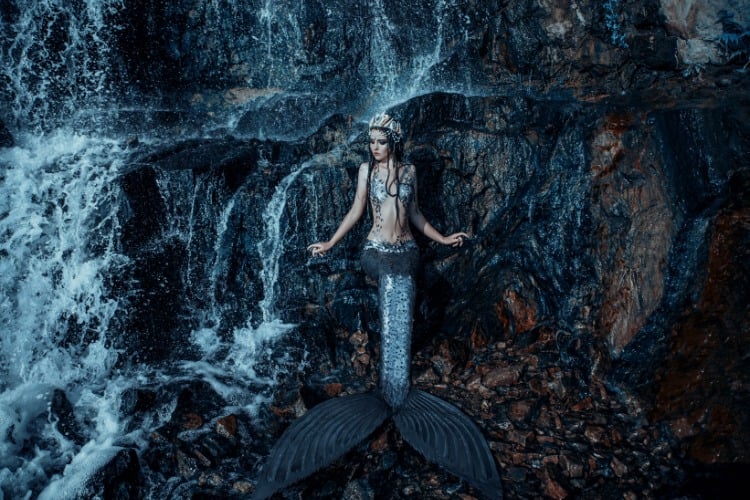 Foto de mulher vestida de sereia sentada em pedras de local Com parede de pedra Possivelmente uma cachoeira com água caindo