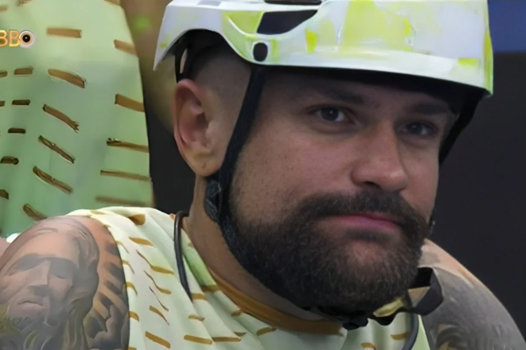 foto mostra Vinícius Rodrigues do BBB 24 durante a Prova do Líder. Rapaz está com uma expressão séria, vestindo um capacete de cor branca