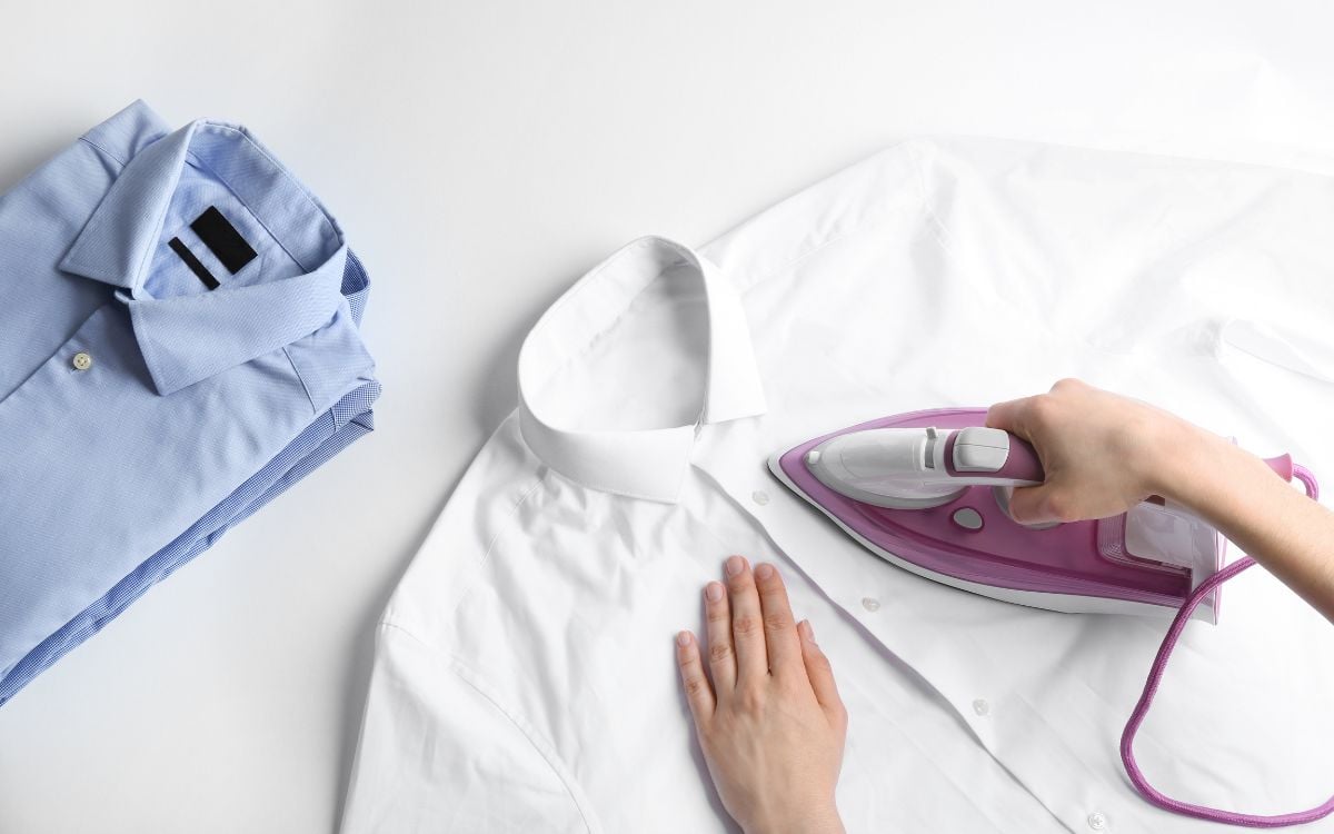 Mãos de mulher segurando ferro de passar roupa profissional em cima de camisa branca. Ao lado, vemos uma pilha de camisas azuis dobradas