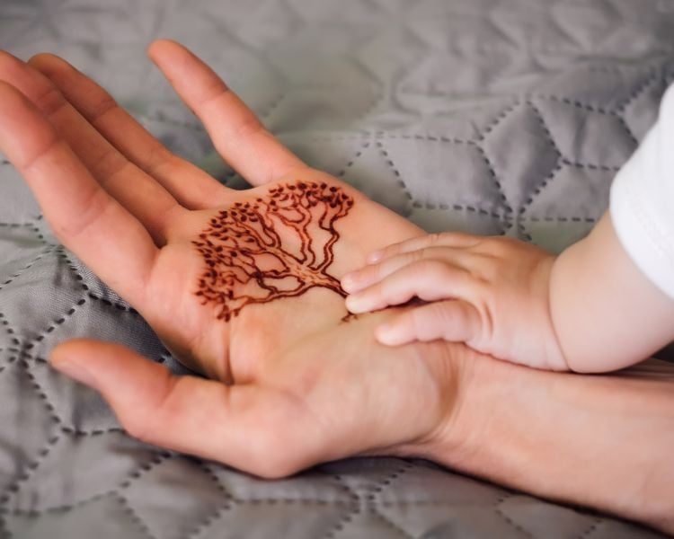 Mão de adulto com árvore genealógica de henna e mão de bebê em cima, ambas sobre um cobertor cinza