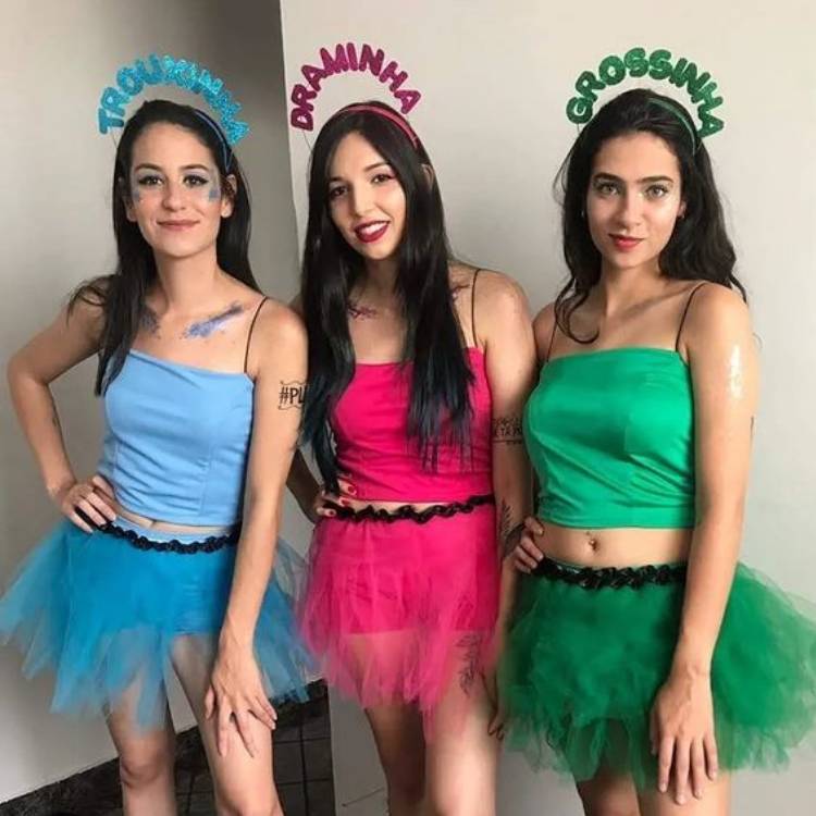 Meninas de pele clara e cabelo escuro usando roupas azul, rosa e verde, inspiradas nas meninas Super Poderosas, com tiaras com escrito "grossinha", "trouxinha" e "draminha".