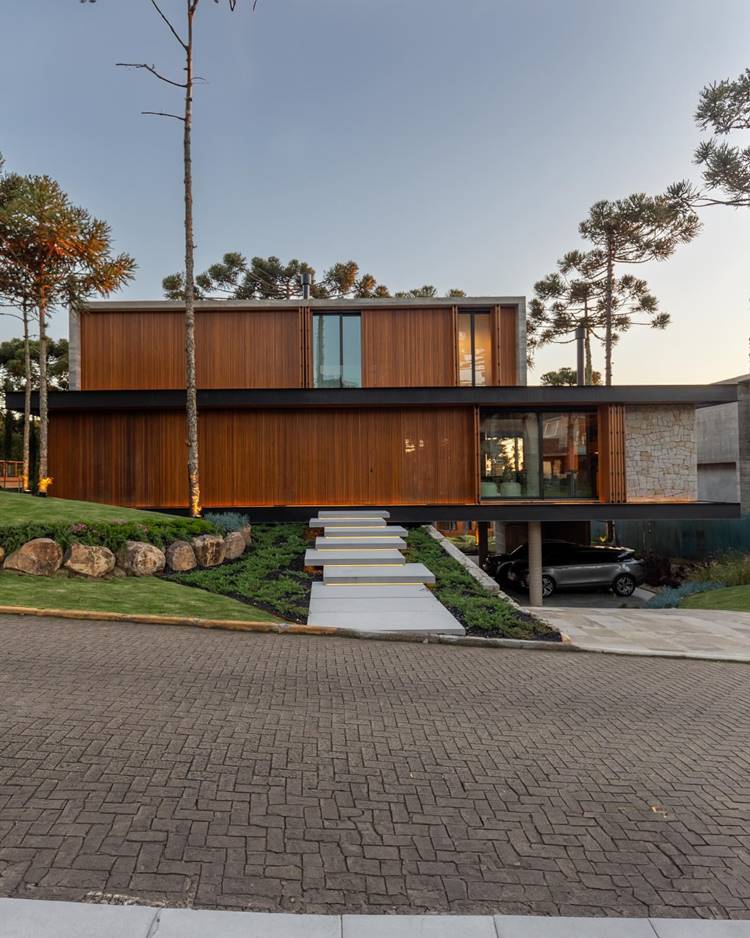 A imagem mostra uma casa moderna, com paredes de madeira e concreto, janelas grandes e está cercada por árvores. A casa tem uma escada branca que leva à entrada, uma garagem aberta com um carro e um céu claro