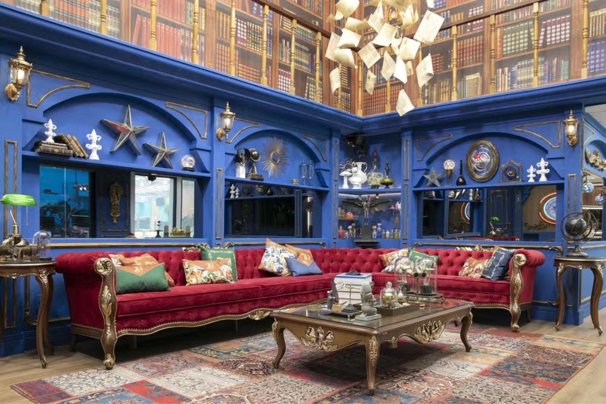 Foto da sala do BBB 24, com sofá vermelho, parede azul, papel de parede de estante de livros, mesa de centro de madeira, almofadas e diversos itens decorativos.