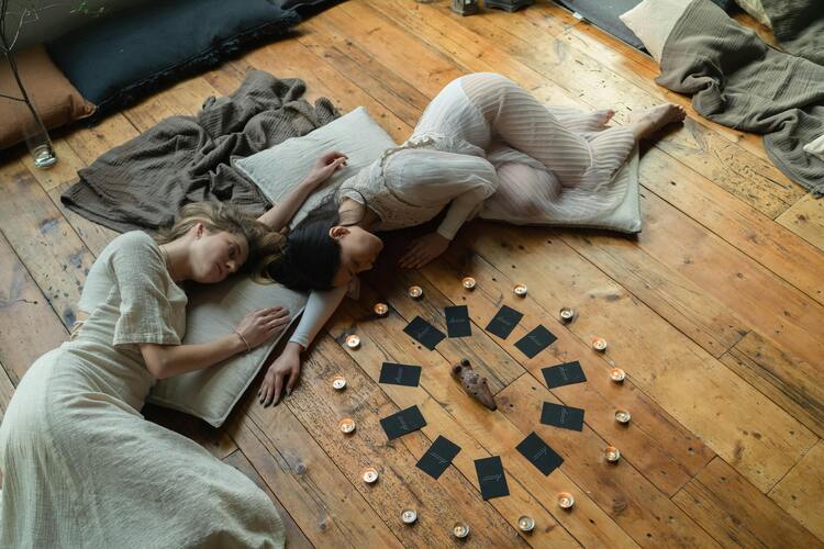 Duas mulheres deitadas no chão como se estivessem fazendo uma magia, até mesmo uma amarração amorosa. Ao lado, é possível ver cartas de tarot e outros elementos mágicos