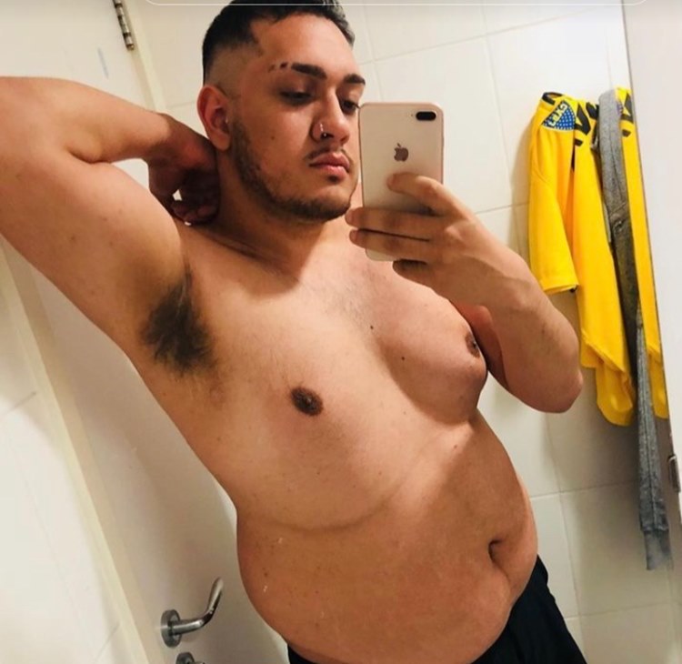 Foto antiga do funkeiro do BBB 24 sem camisa em uma selfie no espelho, usando bermuda preta e exibindo um de seus braços. O banheiro possui um suporte com uma blusa amarela e outra peça de roupa pendurada.