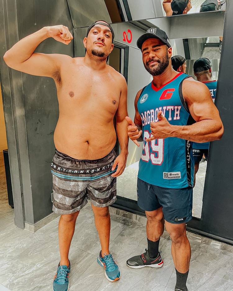 Foto do funkeiro do BBB 24 sem camisa, usando uma bermuda estampada e tênis, ao lado de um profissional de educação física musculoso que veste regata, shorts, tênis e meia comprida, além de um boné. Ambos estão em frente a um espelho em uma parede preta, possivelmente em uma academia.