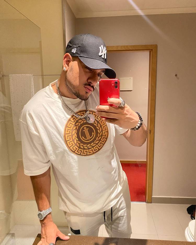 Mc Bin Laden após emagrecer, em uma selfie com um iPhone vermelho, no espelho de um banheiro. Ele está vestindo uma camisa branca com estampa de mandala marrom e bege, uma calça branca de moletom, joias pratas e um boné preto da New York.