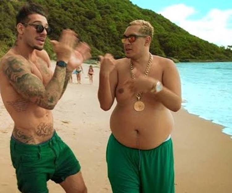 Print de Mc Bin Laden no clipe "Tá Tranquilo Tá Favorável", sem camisa, usando cordão dourado e bermuda verde ao lado de outro rapaz, também usando bermuda verde, em uma praia com montanhas ao fundo.