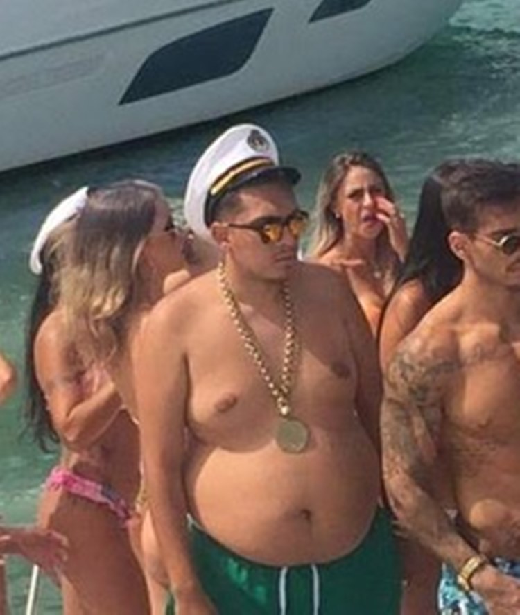 Foto de Mc Bin Laden sem camisa antes de emagrecer, usando cordão dourado, bermuda verde, boina de marinheiro, óculos de sol, rodeado de mulheres de biquínis e também de um homem. Ele está em uma embarcação em alto mar, ao lado de outra embarcação.