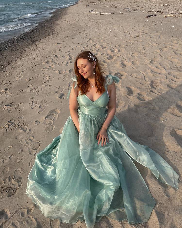 Mulher usando vestido longo verde e penteado com Conchas e estrelas sentada na areia de praia
