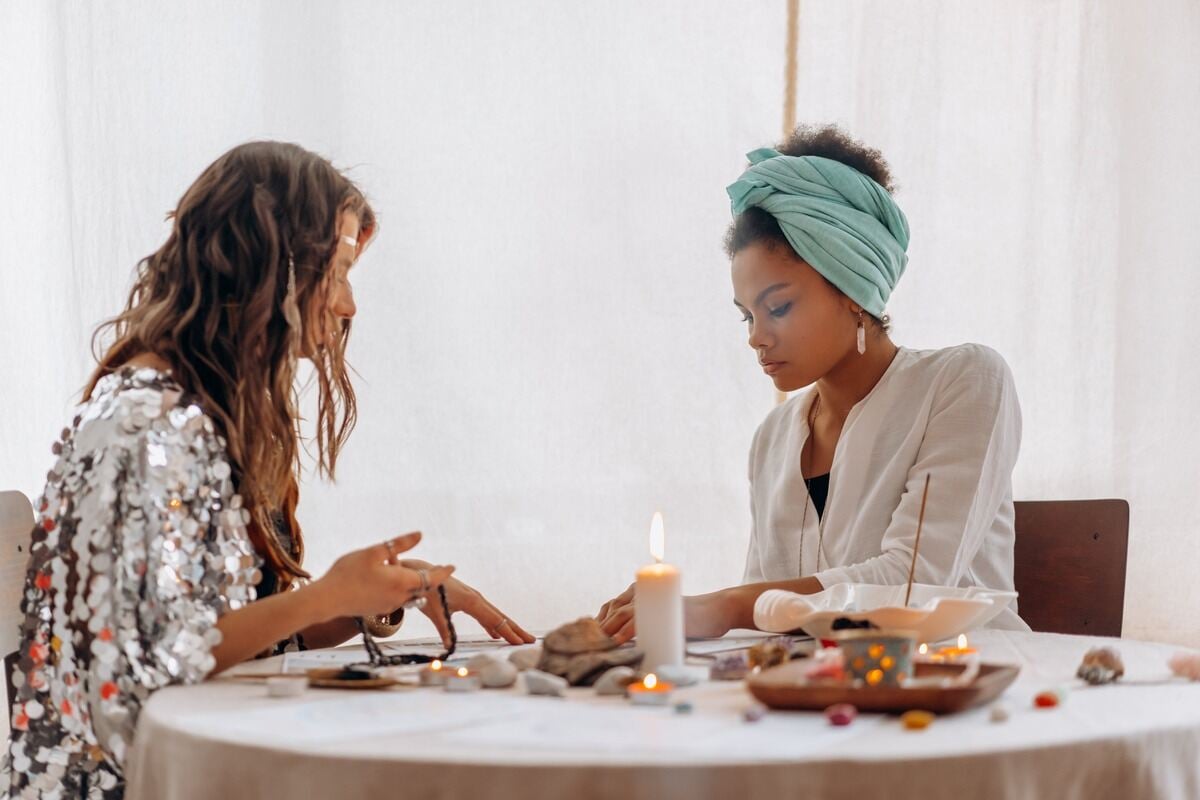 Duas mulheres em uma mesa com velas e uma dela lendo algo, com se fosse uma consulta de terapia holística ou mesa radiônica