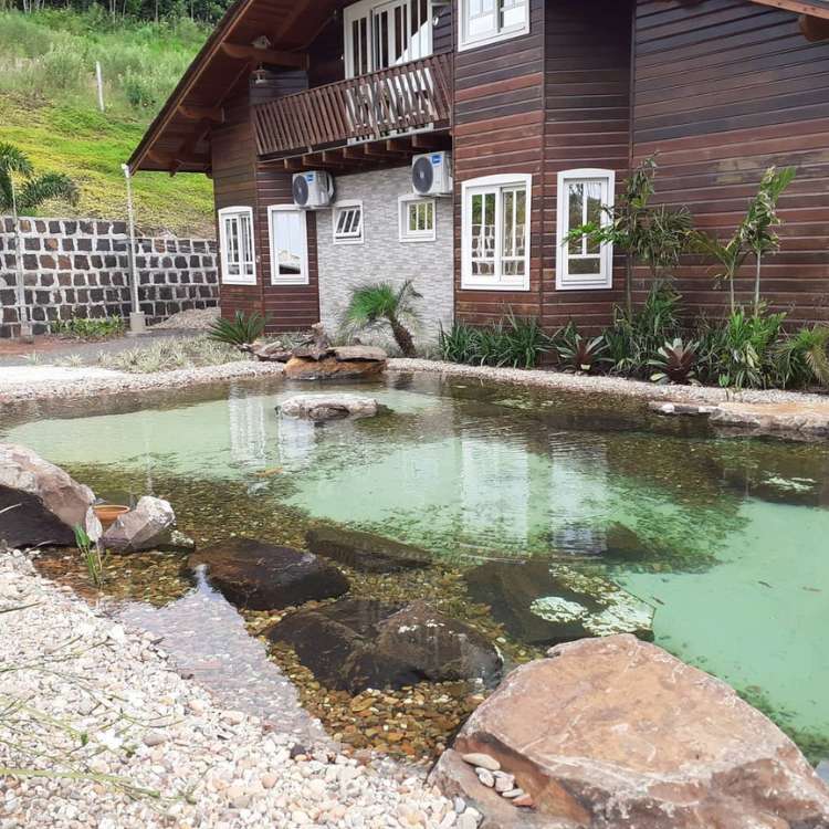 Casa marrom e cinza com piscina biológica na frente, com plantas, pedras e água cristalina. 