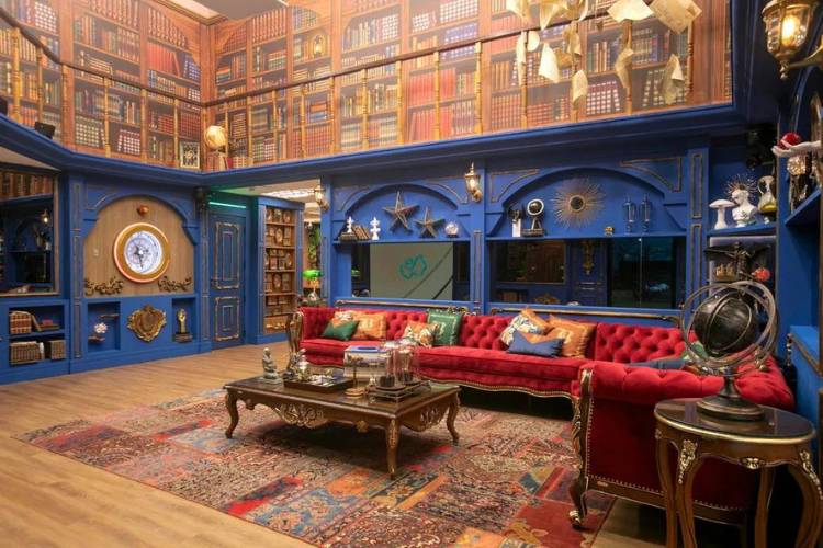 Sala do BBB 24 com papel de parede de biblioteca, parede azul, sofá vermelho, relógio e outros itens decorativos. 