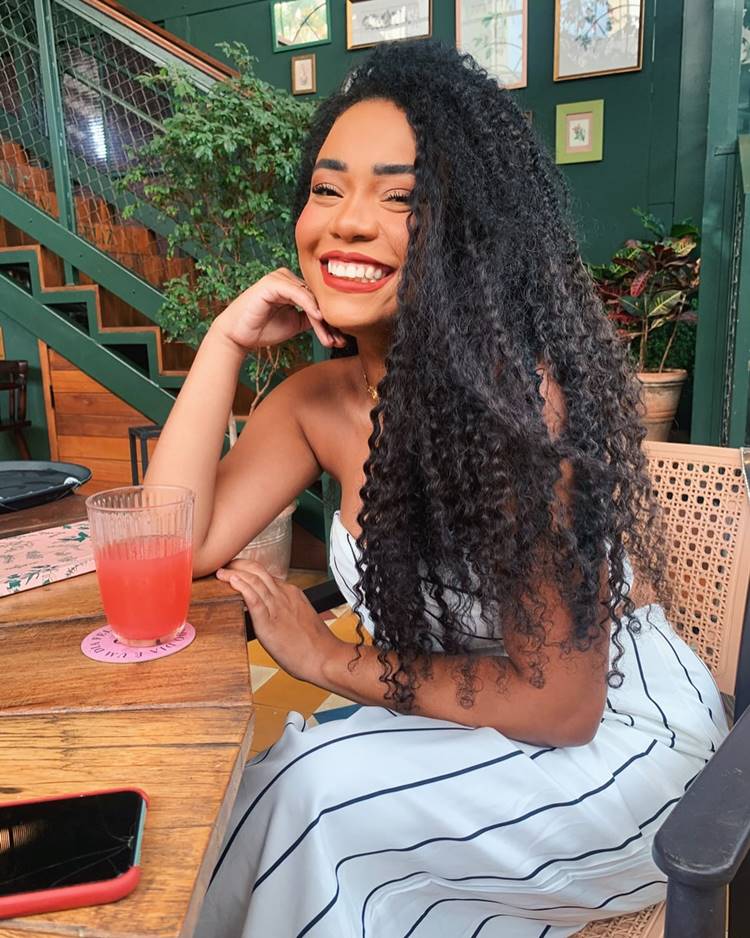 Thalyta Alves do BBB 24 sentada em cadeira em mesa com um copo de refresco vermelho