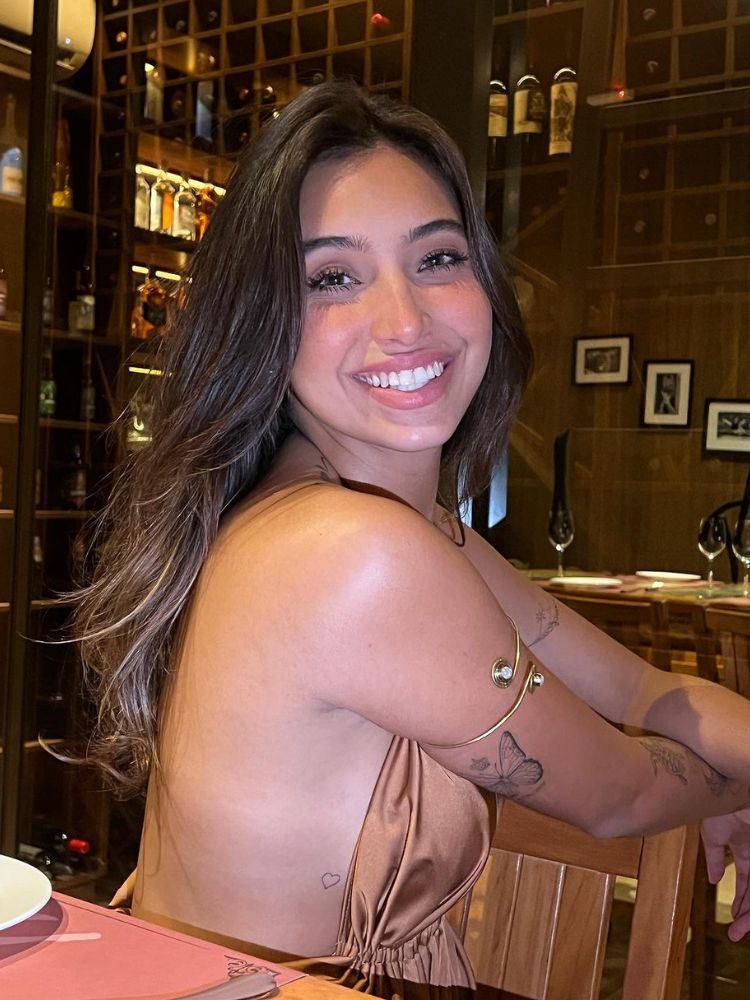 foto de Vanessa Lopes sentada em uma cadeira de madeira de um restaurante. Ela sorri, usando blusa de cetim de frente única e bracelete dourado