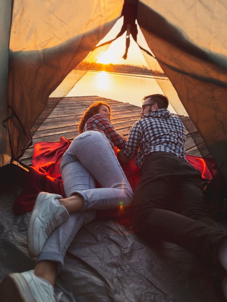 Casal deitados em barraca acampando na beira de lago no pôr-do-sol