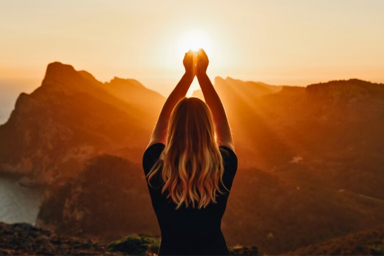 Mulher com as mãos na direação da luz do sol, meditando em meio a montanhas