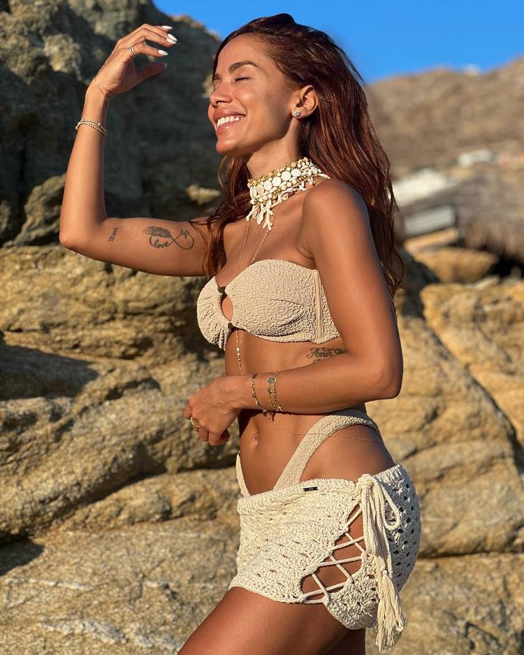 A cantora usando roupa de banho nude com minissaia de crochê e colar de contas na praia, com fundo de pedras.
