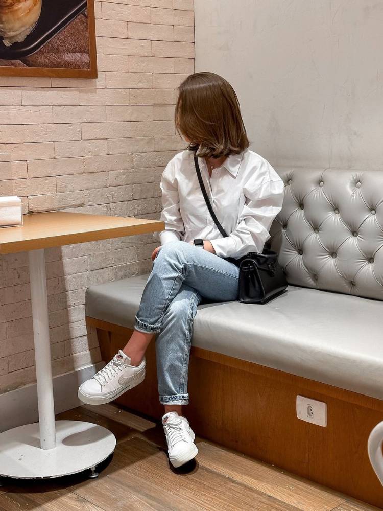 Mulher de rosto virado sentada em sofá de restaurante usando camisa branca, calça jeans curta e tênis branco