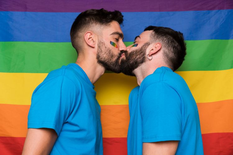 Casal de dois homens com blusa azul se beijando em frente à bandeira LGBT