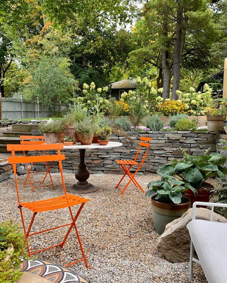 Jardim com pedras, mesa, cadeiras laranjas, plantas e outras flores.