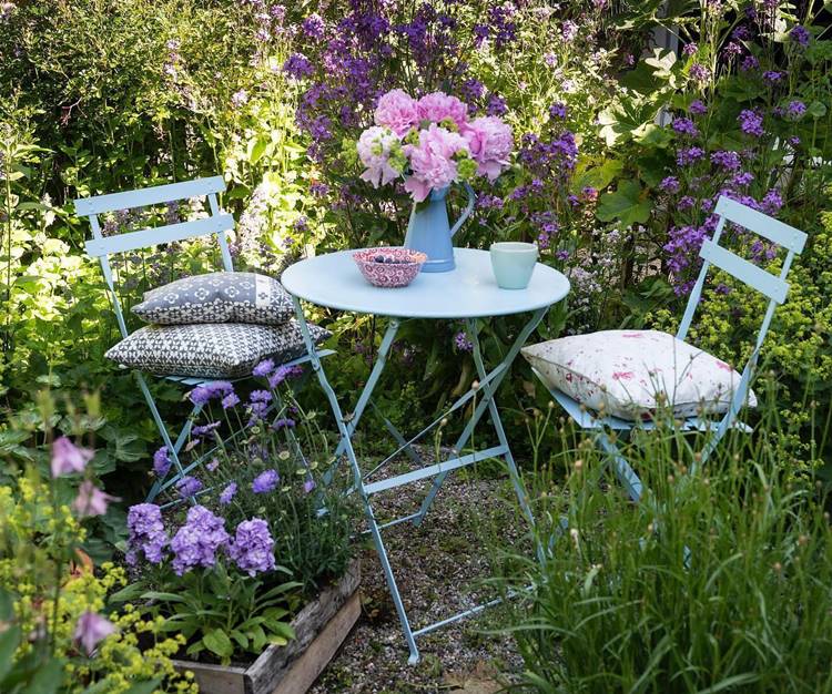 Mesa de ferro azul com almofadas, vaso de flor e xícara em jardim com plantas e chão de pedrinhas.