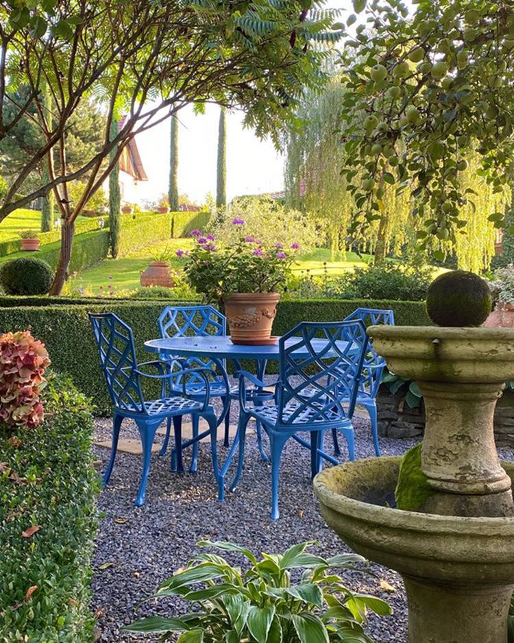 Mesa de ferro azul com vaso de barro com flor em jardim, colchão de pedra e arbustos.