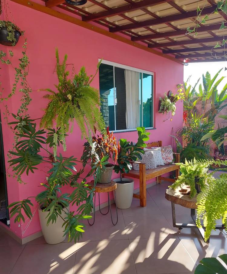 Varanda de casa rosa com muitas plantas, banco de madeira e mesa de madeira.