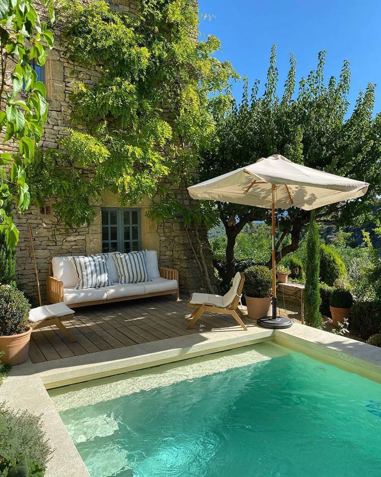 Área de piscina com guarda-sol, cadeiras e sofás, jardim com plantas em vasos e árvores grandes, além de arbustos.