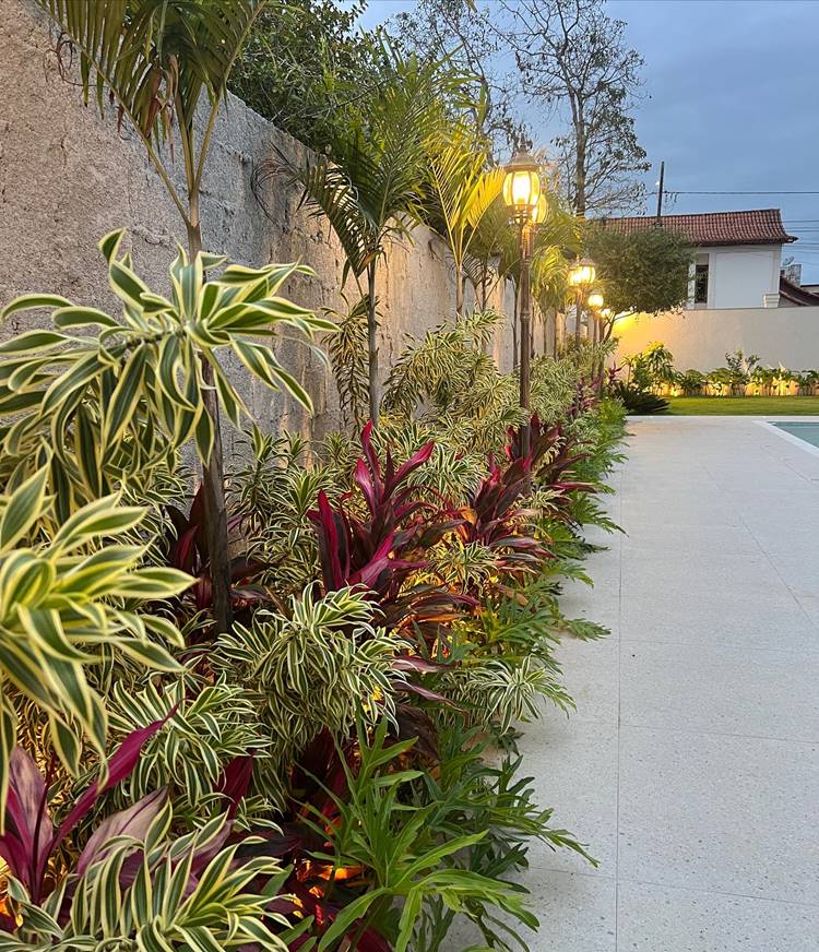 Jardim próximo aos muros com plantas verdes coloridas, como vermelhas e palmeiras, além de luminárias grandes e altas.