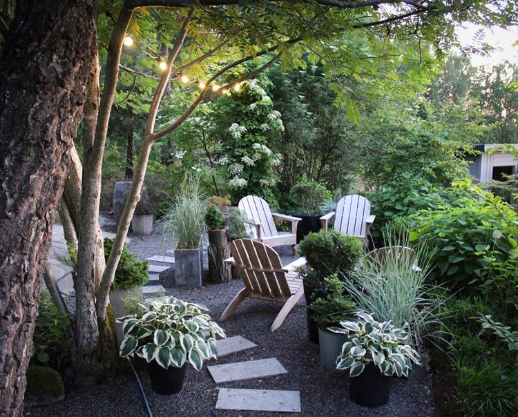 Jardim externo com pedras pequenas no chão, caminho com pedra, plantas em vasos, mesa de madeira, varal de luzes e outras plantas.