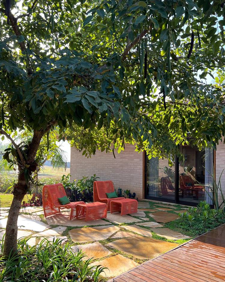 Jardim tropical com chão de pedra e grama, cadeiras e mesas laranjas com almofadas verdes, árvore e outras plantas.