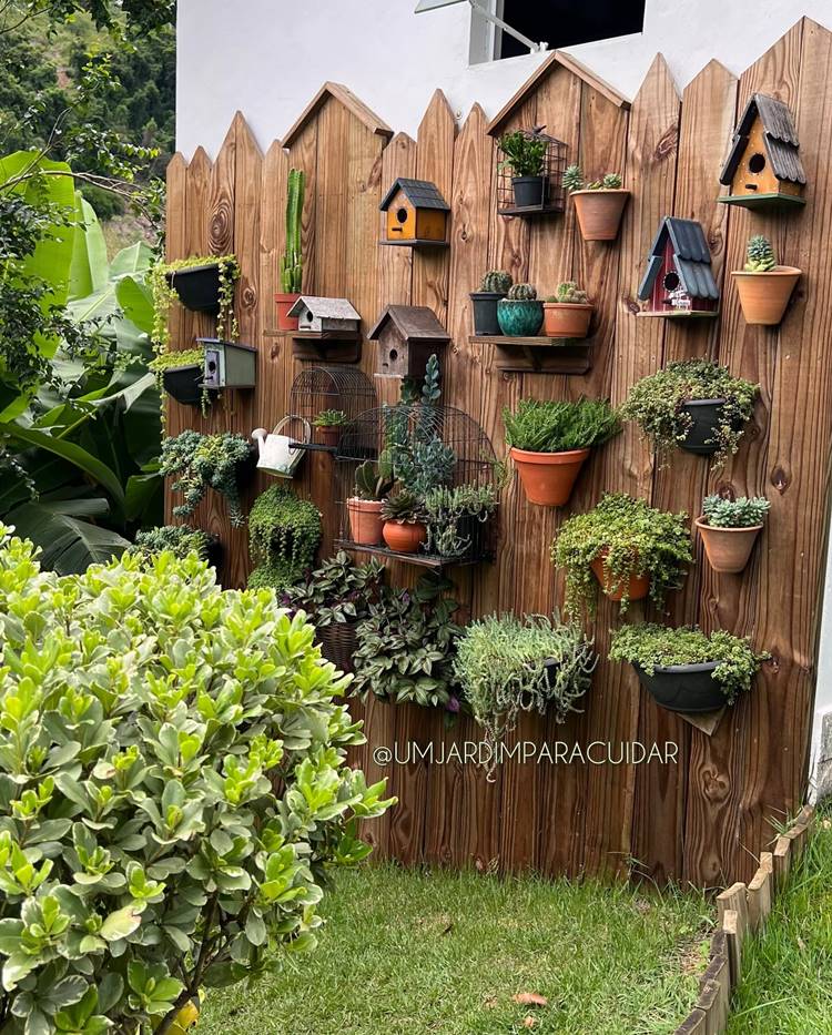 Jardim vertical de madeira com vasos de plantas e casinhas de passarinho ao redor, além de outras plantas como bananeira.