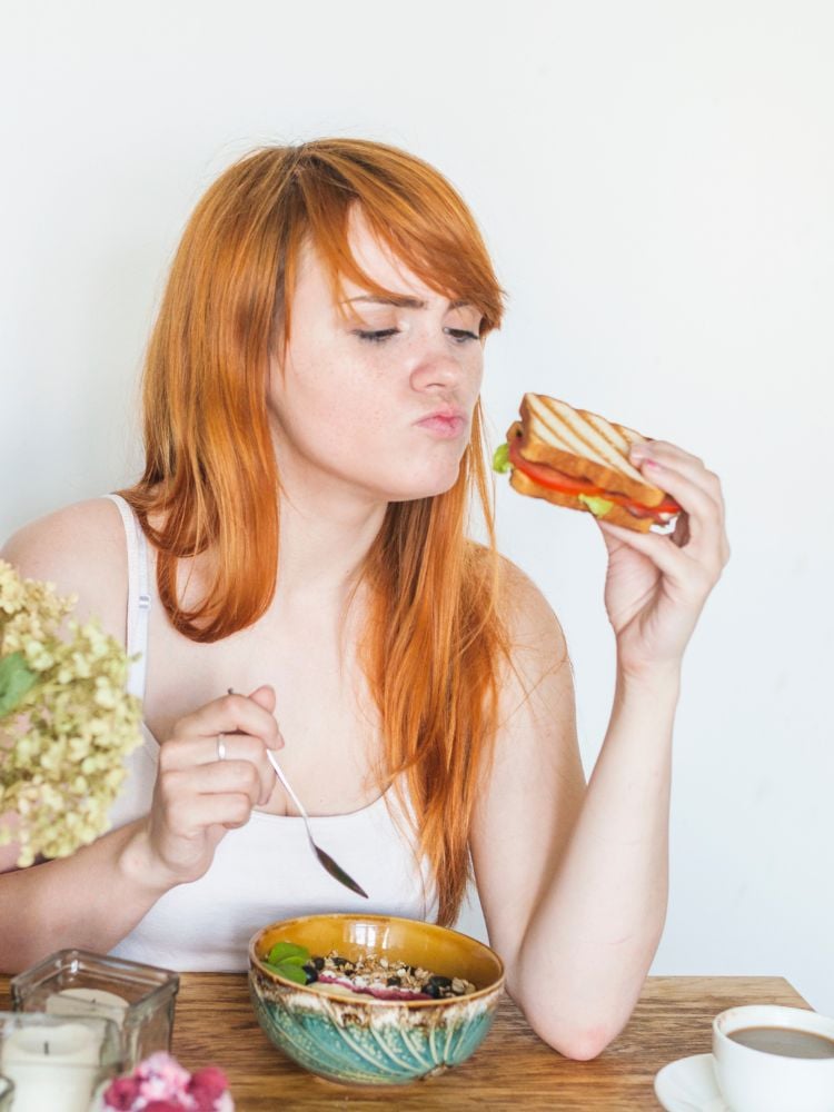 Mulher de pele clara e cabelo ruivo segurando um sanduíche e uma colher, próxima a um pote com comida