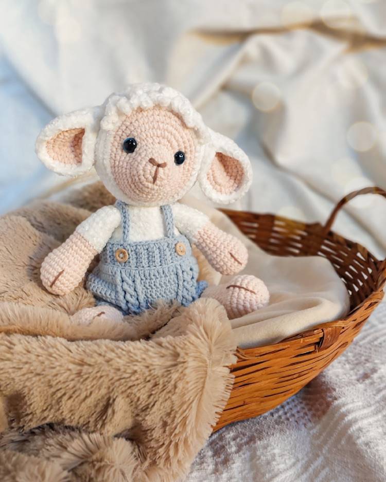 Cordeirinho de crochê branco com roupinha azul, acomodado em uma cesta adornada com tecidos, e mais tecidos ao fundo.

