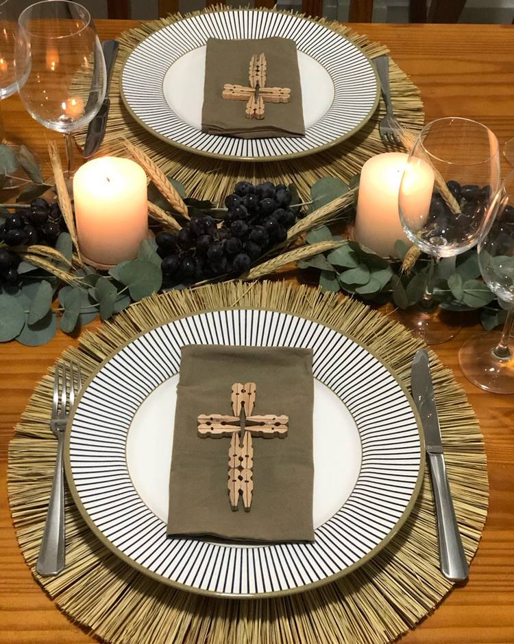 Uma mesa de jantar decorada para a Páscoa Cristã: sousplat de bambu, prato de cerâmica, guardanapo com cruz de madeira decorada em cinza, centro de mesa com flor, uvas, velas e taças ao redor.